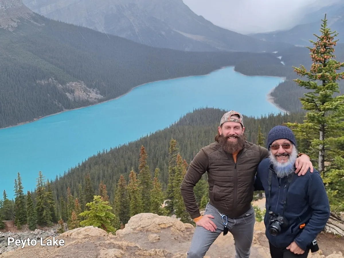 Two men posing with lake and green mountains at the back.