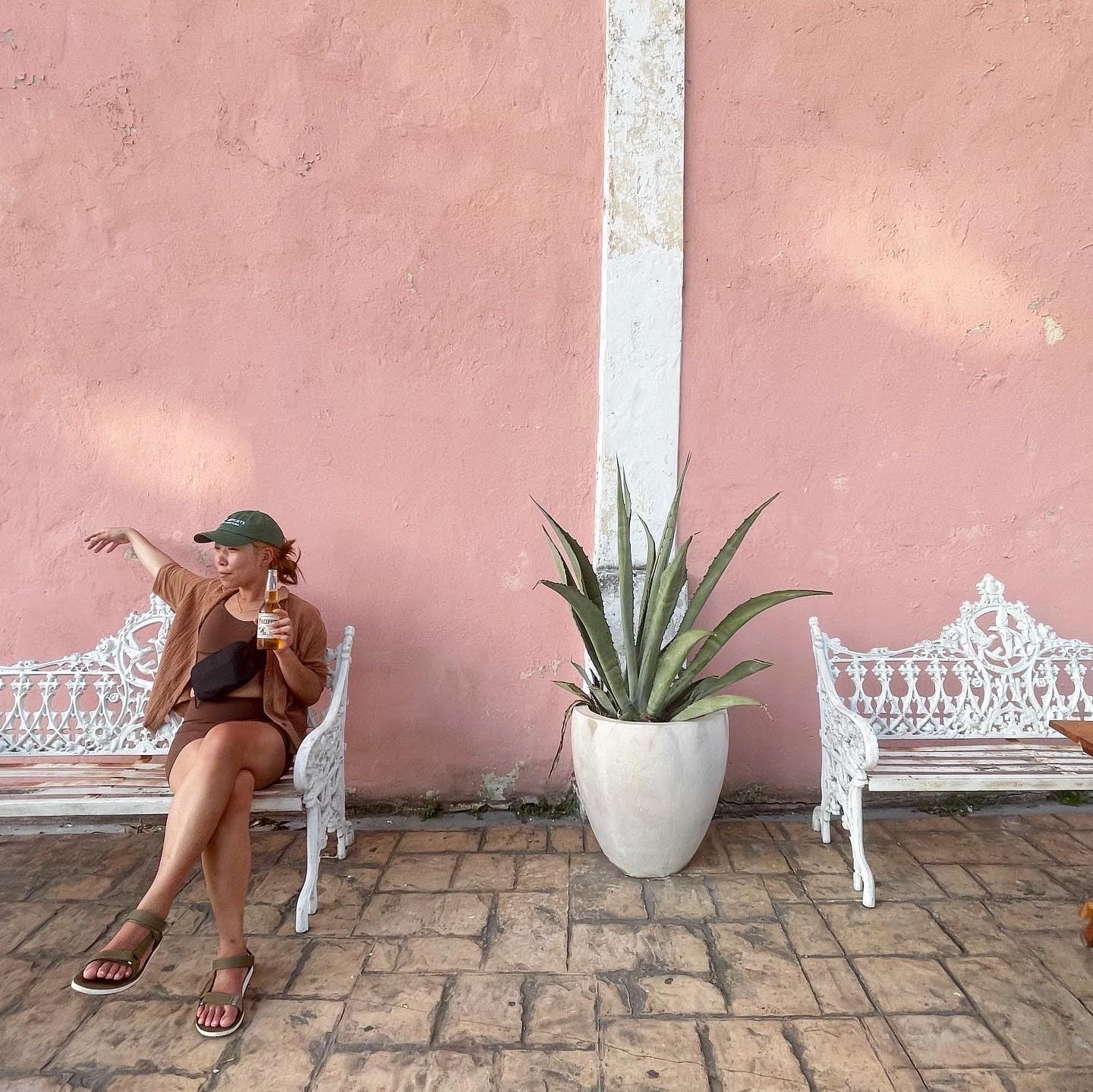 Kimberly sitting on a white bench in front of a pink wall, stone tiling and white pot holding a green plant.