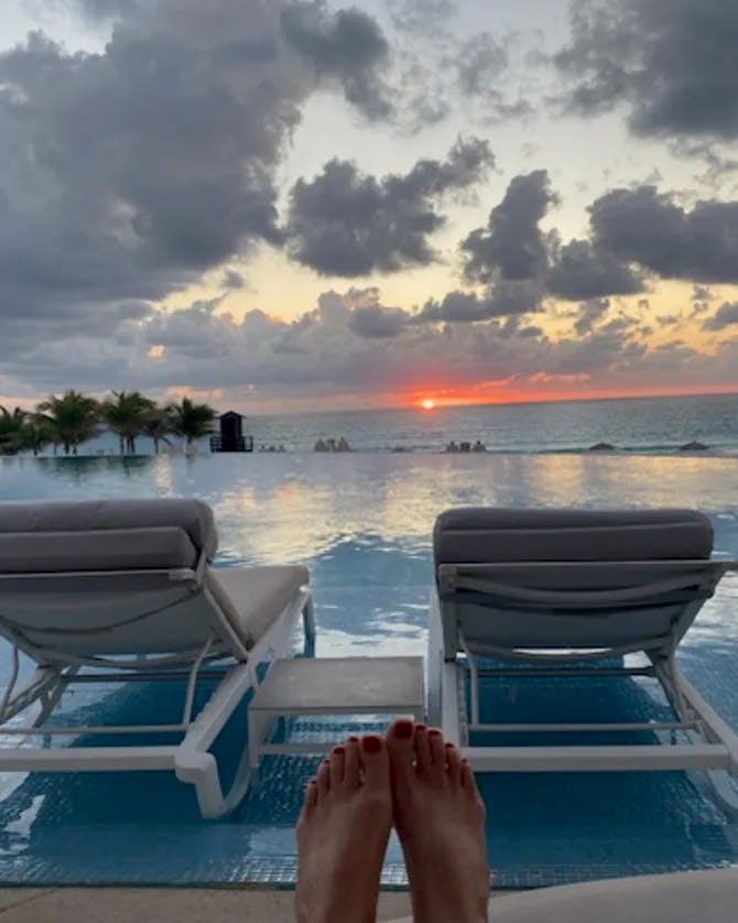 Feet positioned in front of two lawn chairs against a swimming pool that looks out to the sea and a sunset around scattered clouds and palm trees