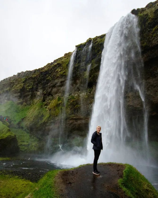 A person standing on a dirt path that leads to a waterfall surrounded by greenery