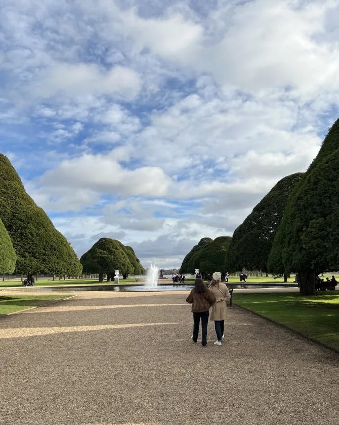 Two people walking down a dirt path surrounded by manicured trees that lead to a fountain