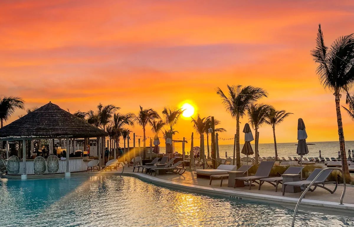 A vibrant orange and pink sunset shining through palm trees and onto a swimming pool deck. 