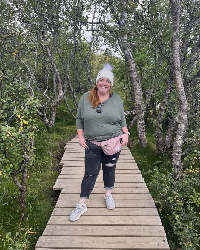 Emyli wearing a white beanie, green top, pink fanny pack and black pants on a wooden pathway surrounded by green trees and bushes