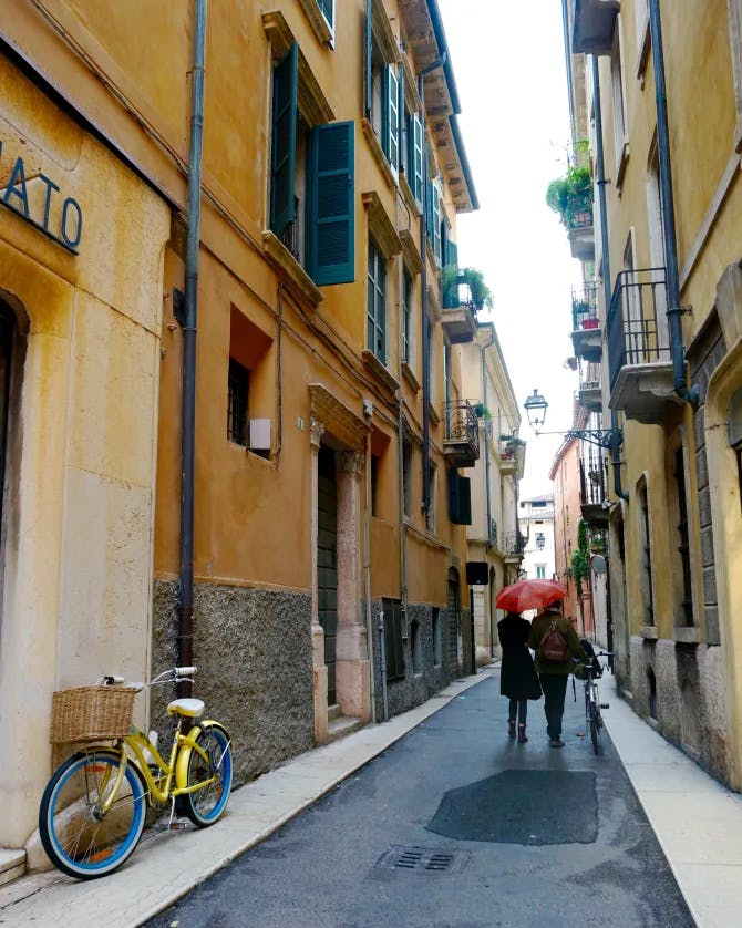 Two people walking down a European city street with a red umbrella in hand, yellow buildings with blue shutters and a bicycle in the surrounding areas