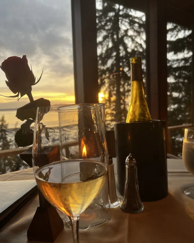 A glass of wine on a dining table in front of a bottle of wine and rose with a sunset and pine trees in the background