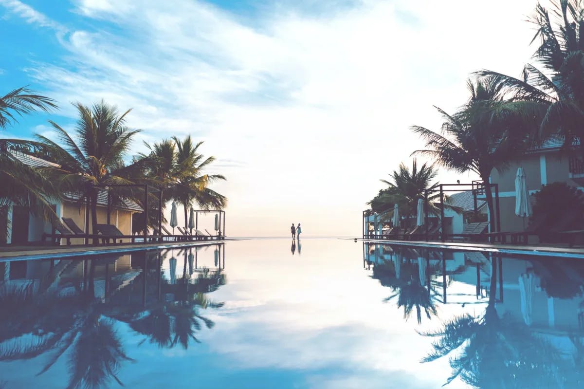 A low-angle photo makes it look like a prominently featured infinity pool blends into the distant ocean with palm trees and cabanas on either side