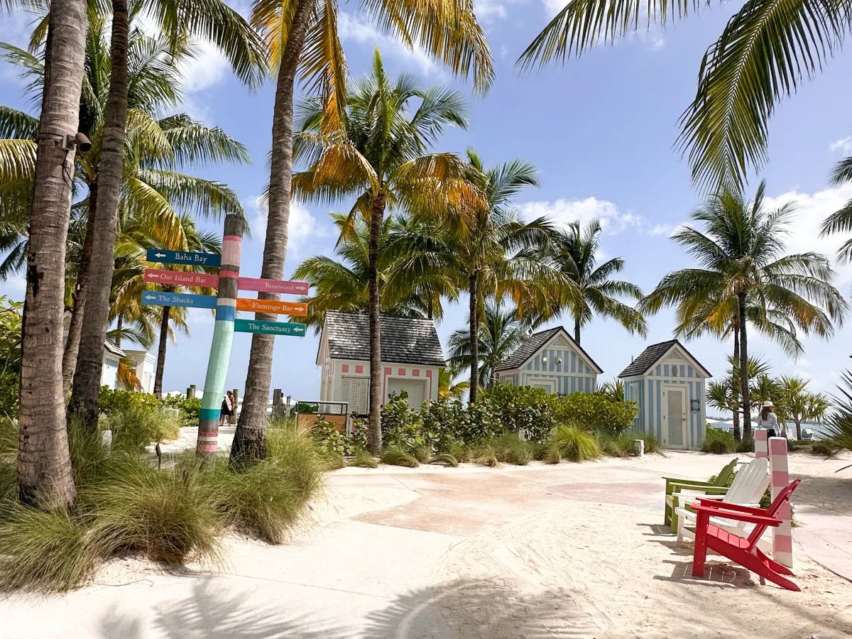A view of a sandy white pathway surrounded by towering palm trees, a red, white and green beach chair, directional signs and three blue, white and pink striped beach shacks in the background. 