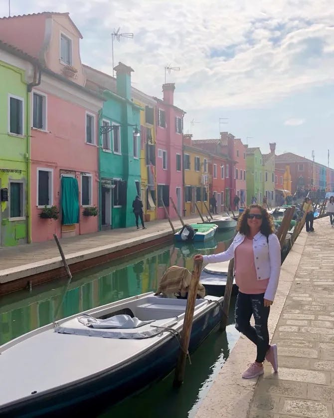 Picture of Katie near boat at Murano with green, pink and blue buildings lining a river and stone path