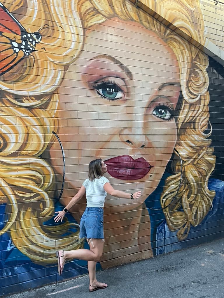 A woman posing in front of a wall painted with a woman's face.