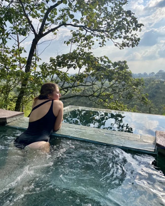 Woman in a black bathing suit sitting in an infinity pool overlooking a lush valley.