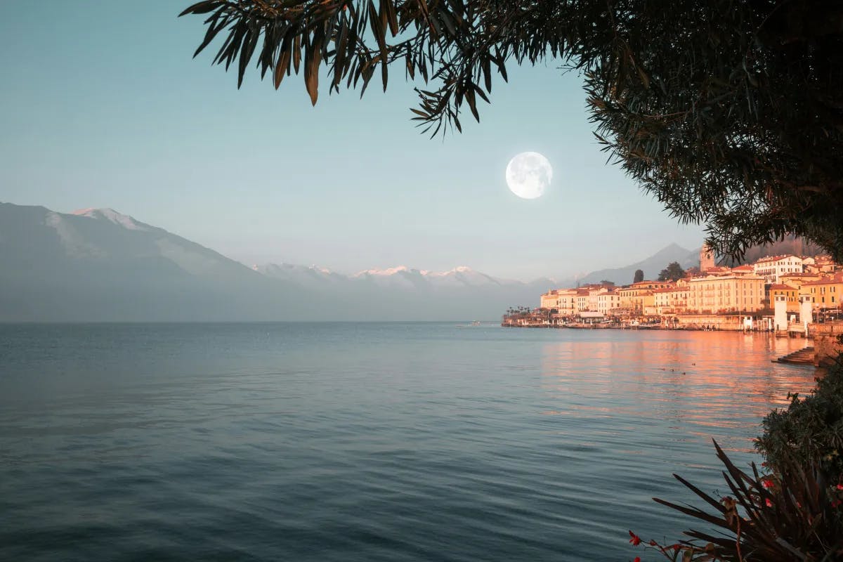A view of the town of Lake Como, Italy next to Lake Como with mountains and a round full moon glowing over the water. 