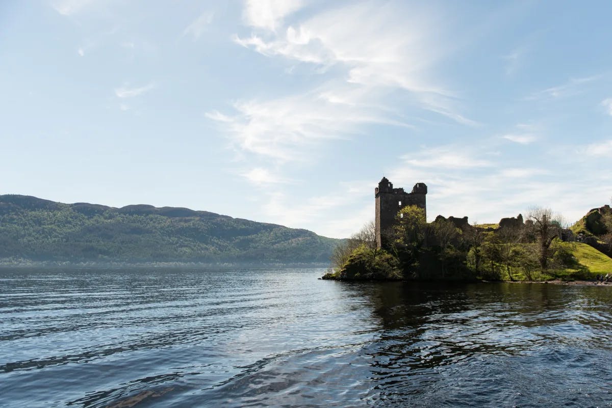 Loch Ness lake in Scotland with blue calm waters and brown castle ruins and on a hill with green grass and more hills in the distance.