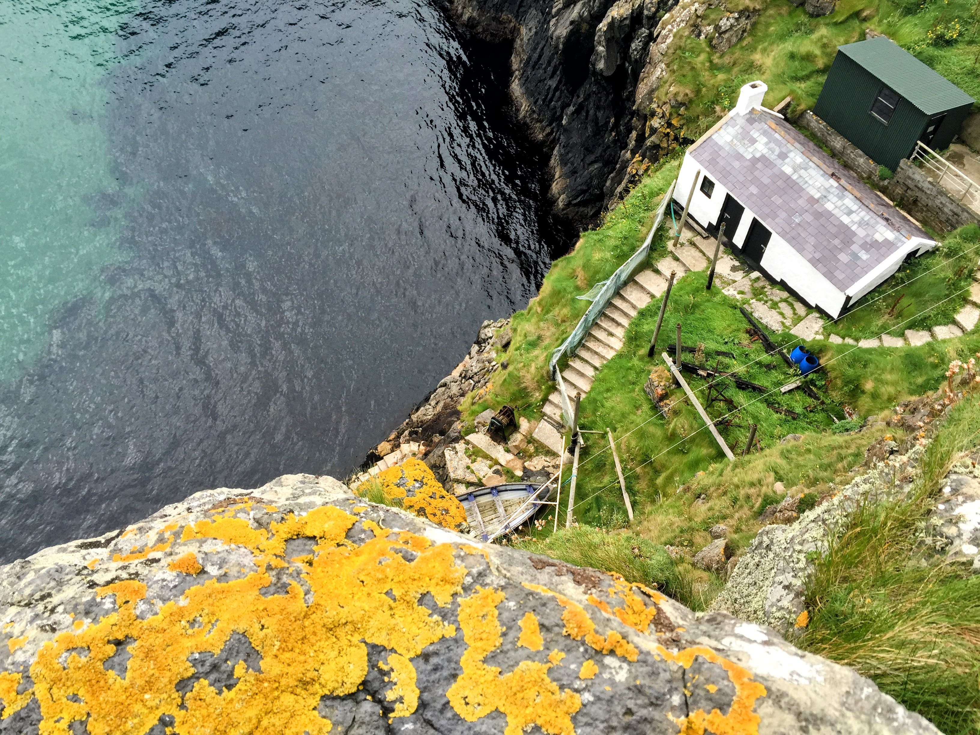 An aerial view of a white house surrounded by green grass with a set of stairs leading to a cliff that overlooks water