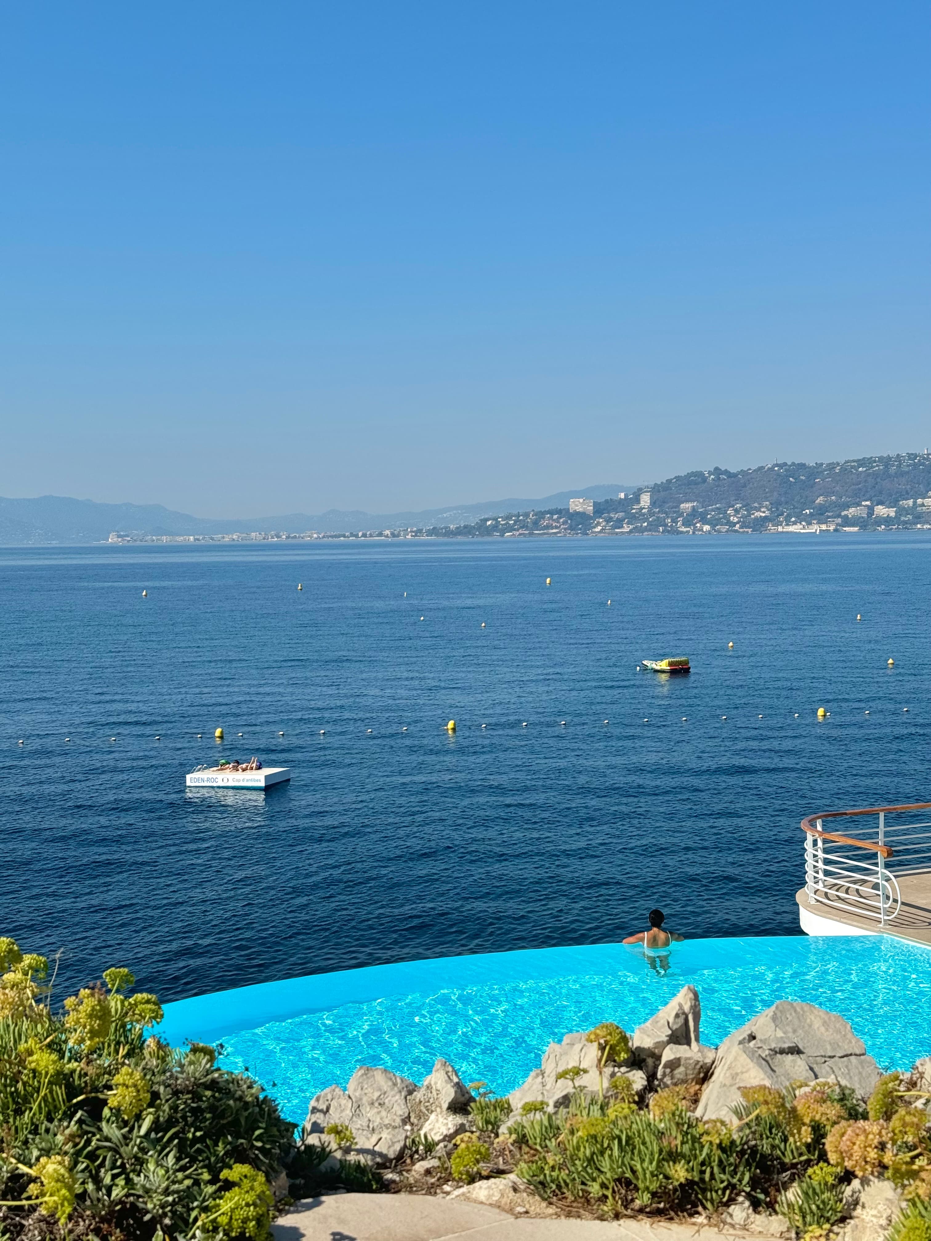 view of an infinity pool, and the sea, with buoys, and a mountainous coastline in the background.