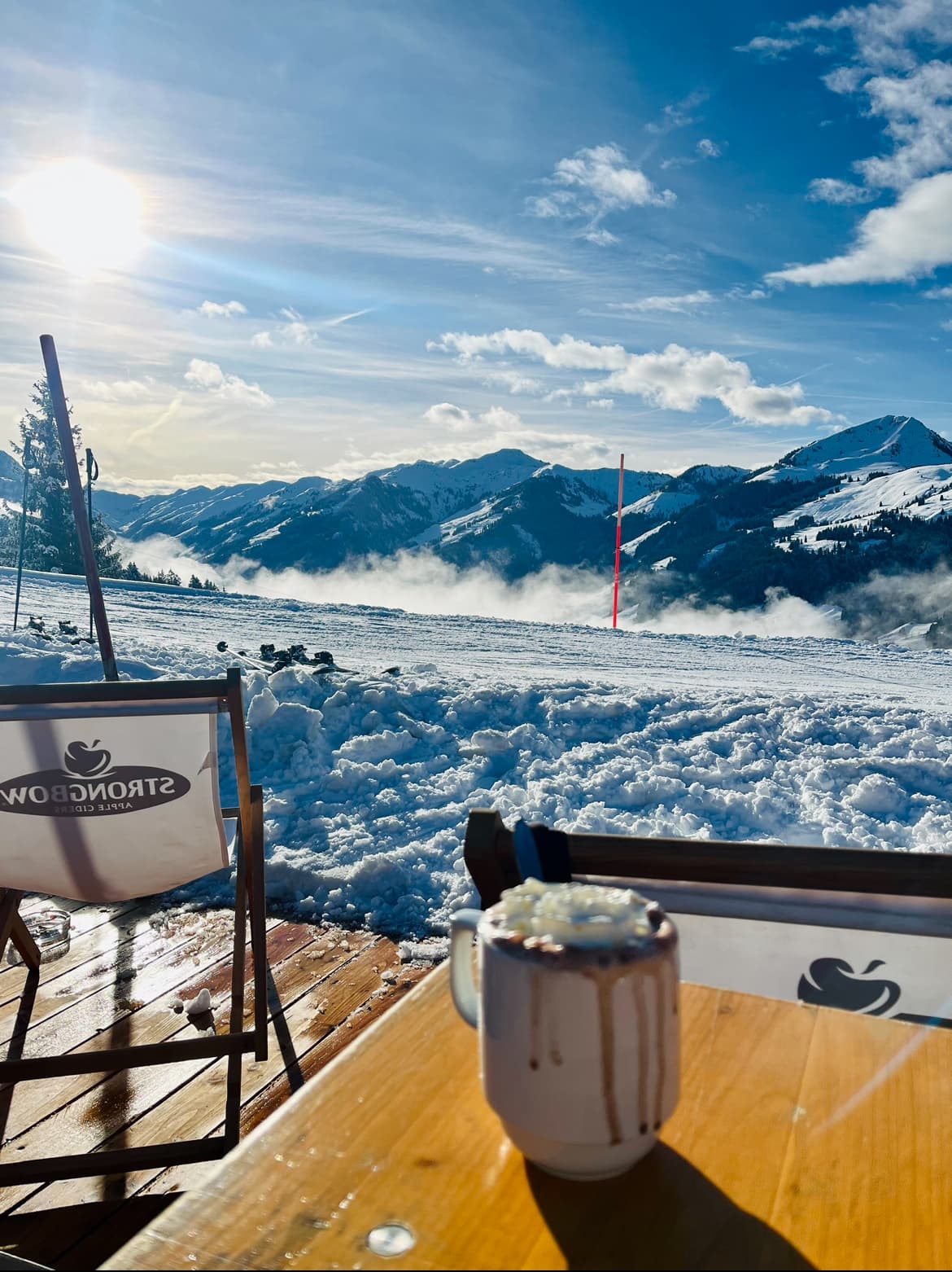 A picture of mountains, thick snow and a mug of hot chocolate on top of a wooden table. There is a deck beneath the table and chair on the left side of the image.