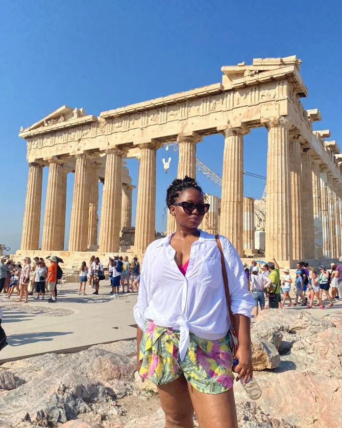 Jia wearing a white top and colorful shorts standing in front of the Parthenon in Greece