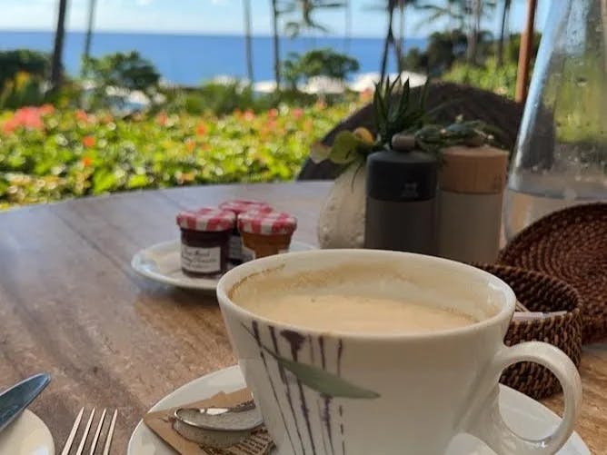 A cup of coffee on a white plate sitting on a table, with a view of the sea