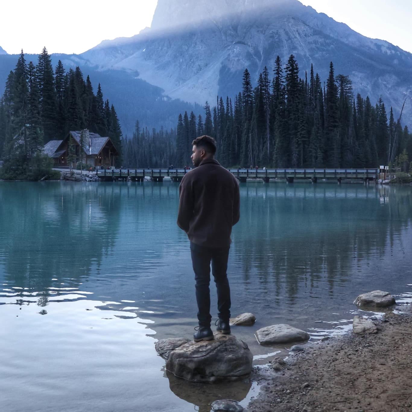 A man wearing a jacket is standing on a rock in lake.