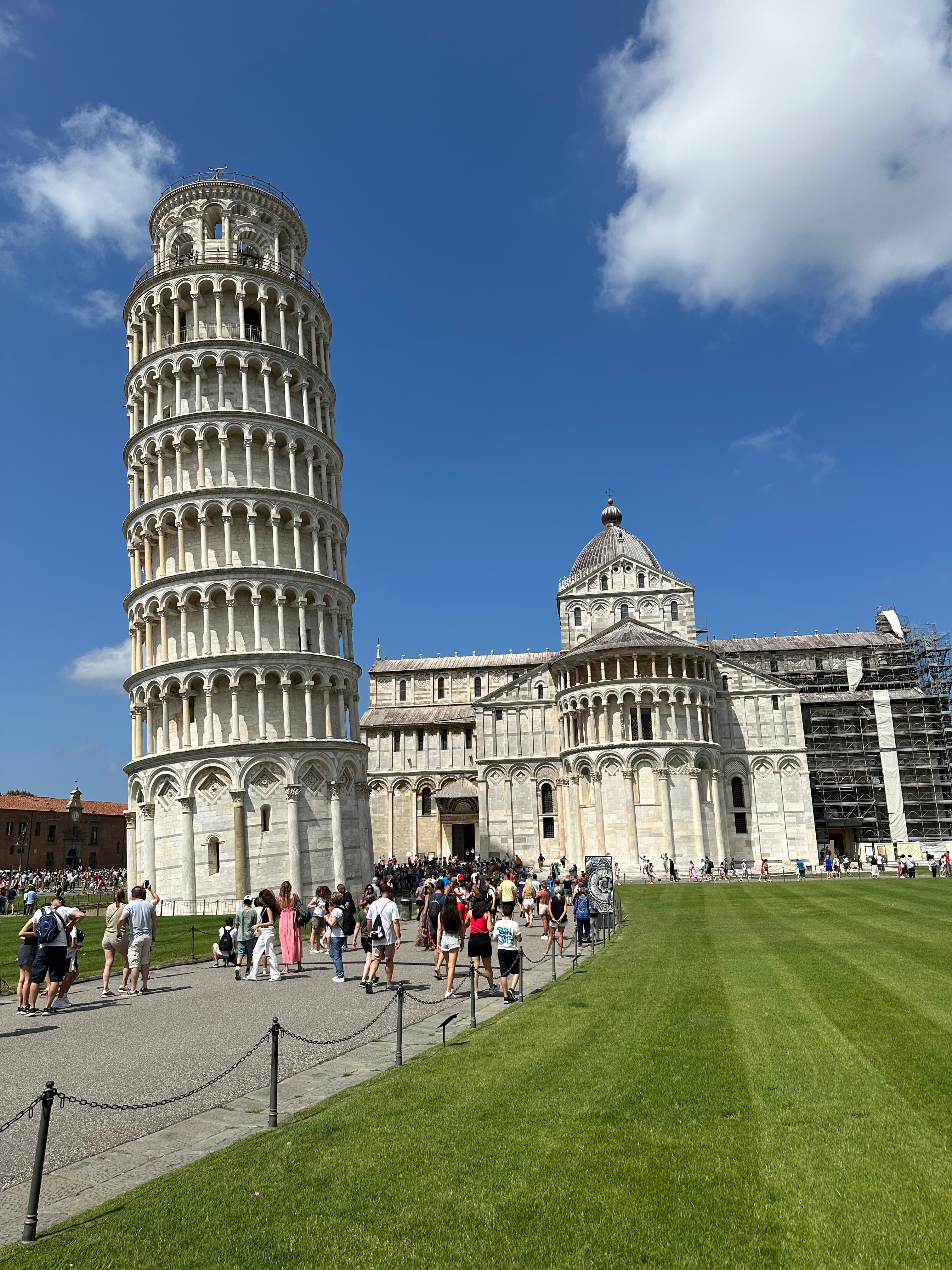 Picture of the Leaning Tower of Pisa against a pathway and field of mowed grass with a church in the background set against the blue sky