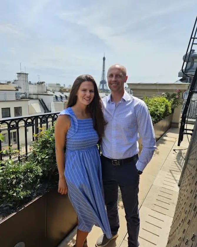 A picture of Meredith wearing a blue dress and standing next to her spouse on a balcony overlooking the Eiffel Tower in Paris, France