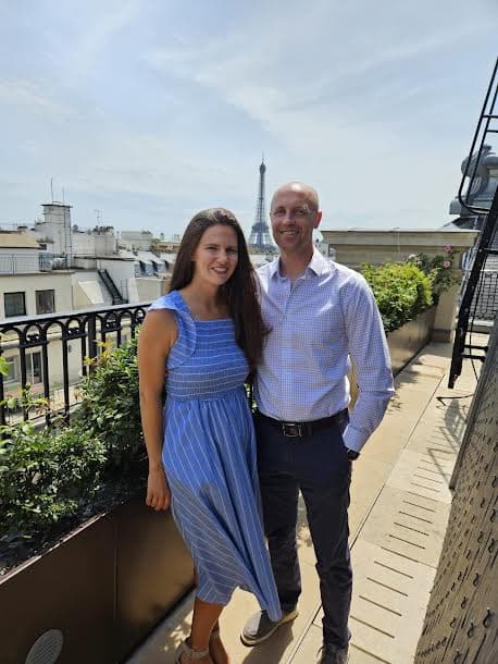 A picture of Meredith wearing a blue dress and standing next to her spouse on a balcony overlooking the Eiffel Tower in Paris, France