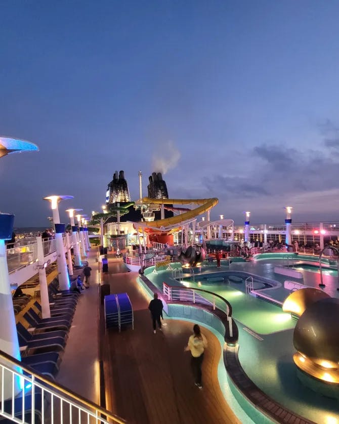 A view of a cruise ship deck with a pool, lawn chairs and colorful lights