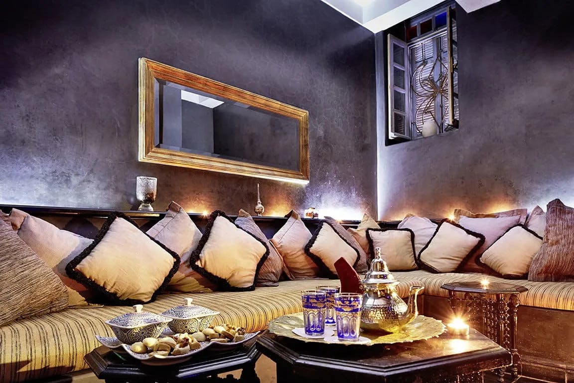 Plush Moroccan pillows fill a massive sofa while a brass tea kettle and pastries sit on a wooden table