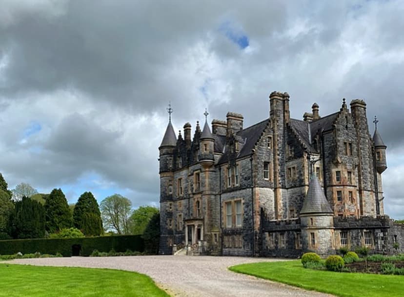 A picture of Blarney Castle & Gardens with a dirt road, green grass and trees surrounding it on a cloudy day