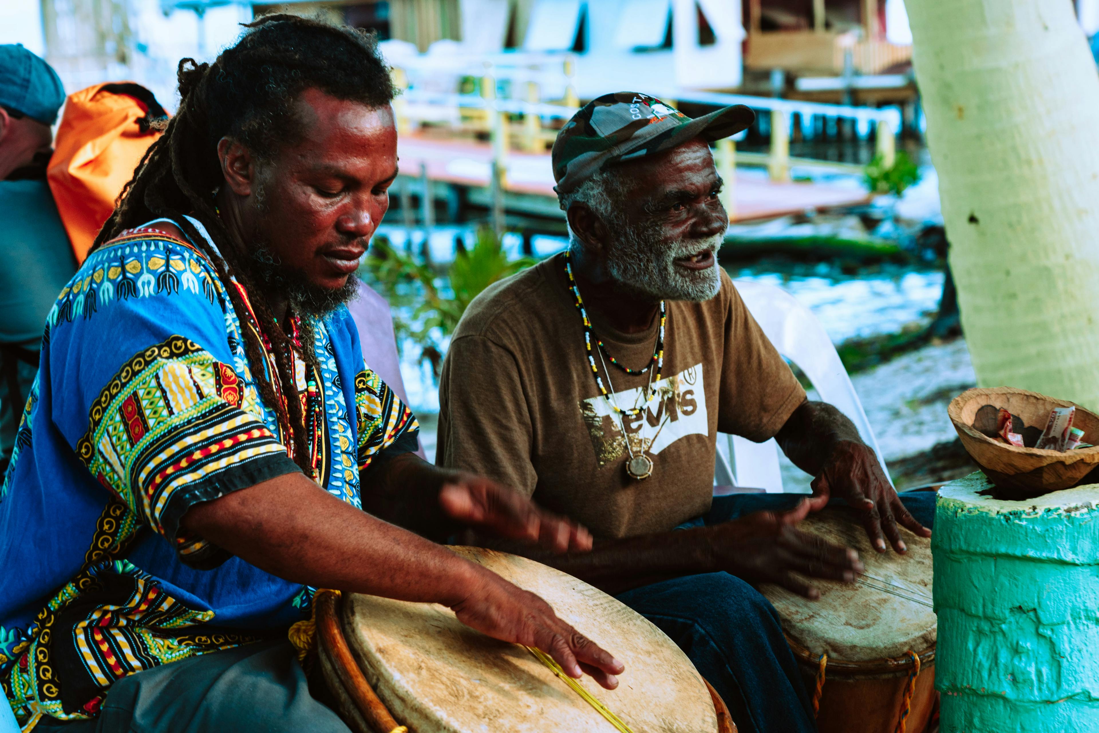 Two men in Belize one with a blue patterned shirt and one with a brown shirt both playing bongo drums
