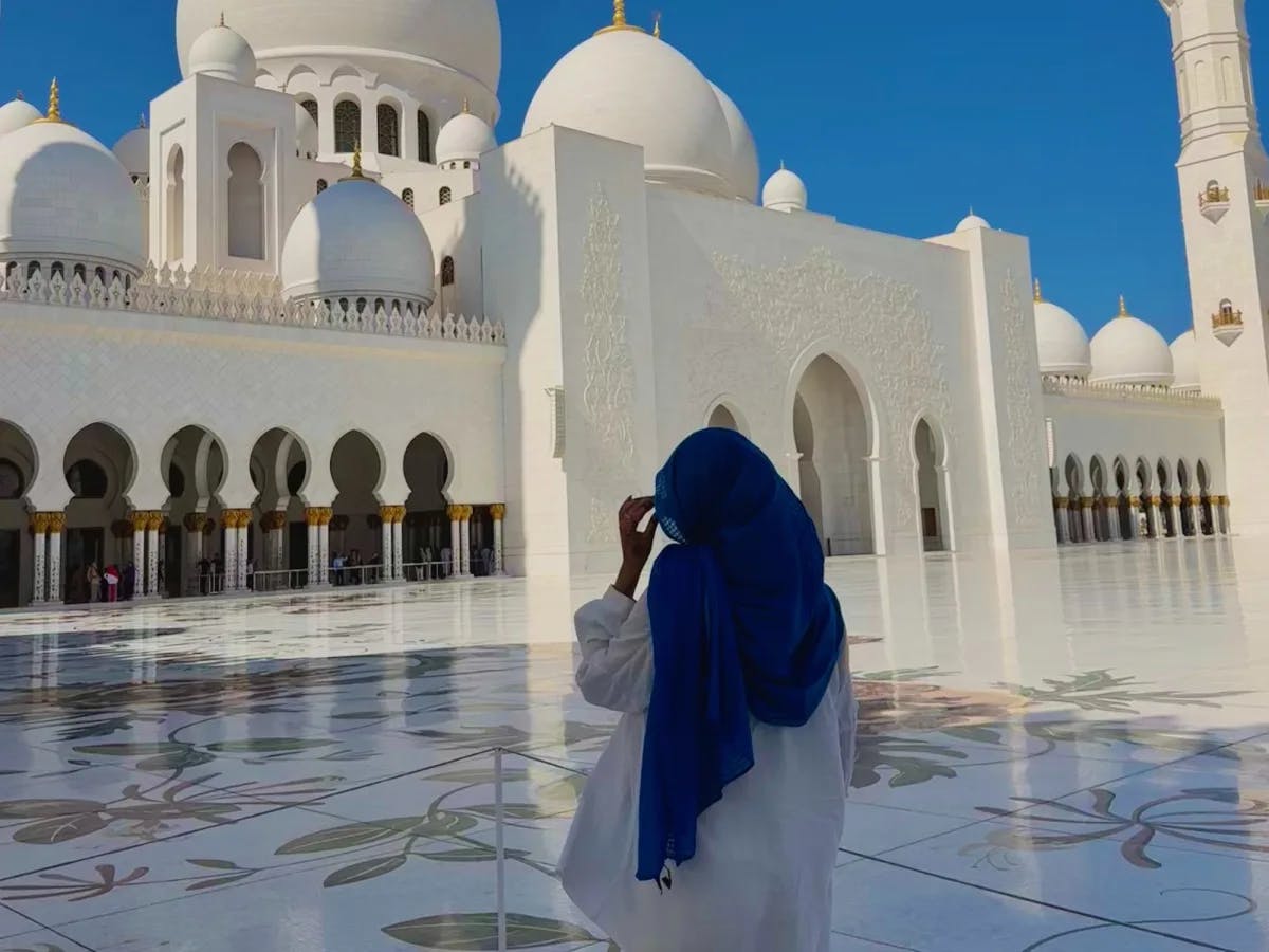 A woman in a blue head covering in front of a white mosque.