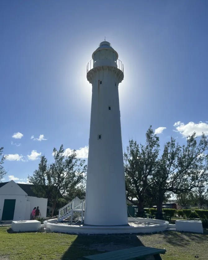 A white lighthouse in front of the bright sun surrounded by trees and grass.
