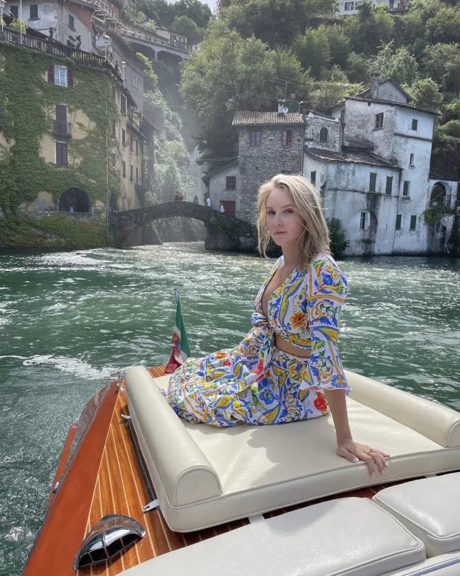 Picture of Alycia wearing a colorful outfit on a boat in front of a bridge, buildings and trees set against Lake Como