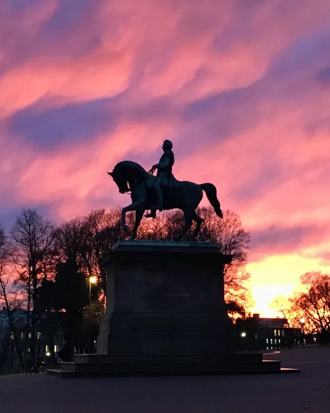 A statue of a man on a horse positioned in front of a vibrant pink and blue sunset.