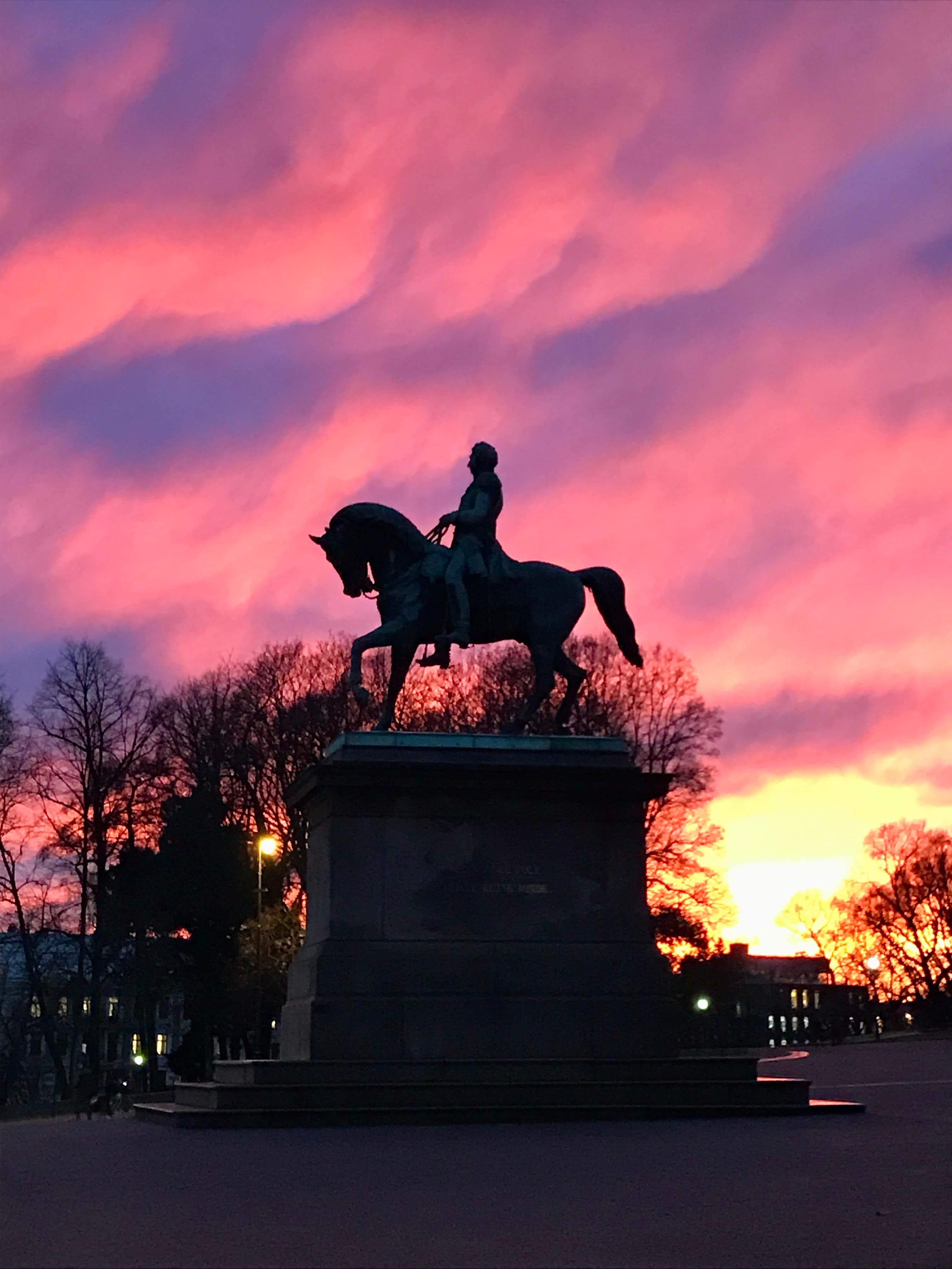 A statue of a man on a horse positioned in front of a vibrant pink and blue sunset.