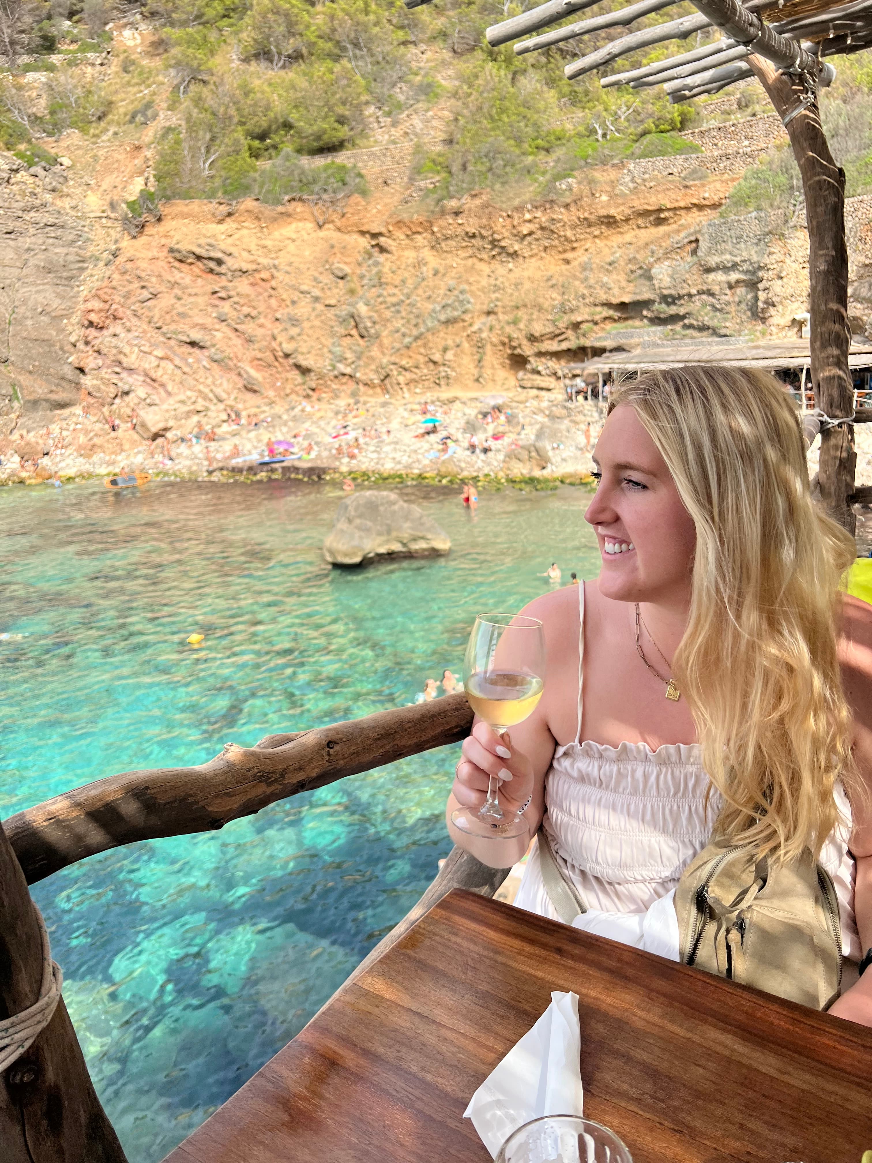 Alyssa wearing a white dress and holding a glass of white wine looking out at the turquoise blue water and rocky terrain