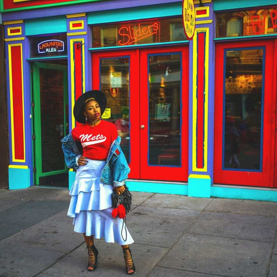 Travel Advisor Azja Greene in a red shirt and light blue skirt with a blue, red, yellow and purple building in the background.