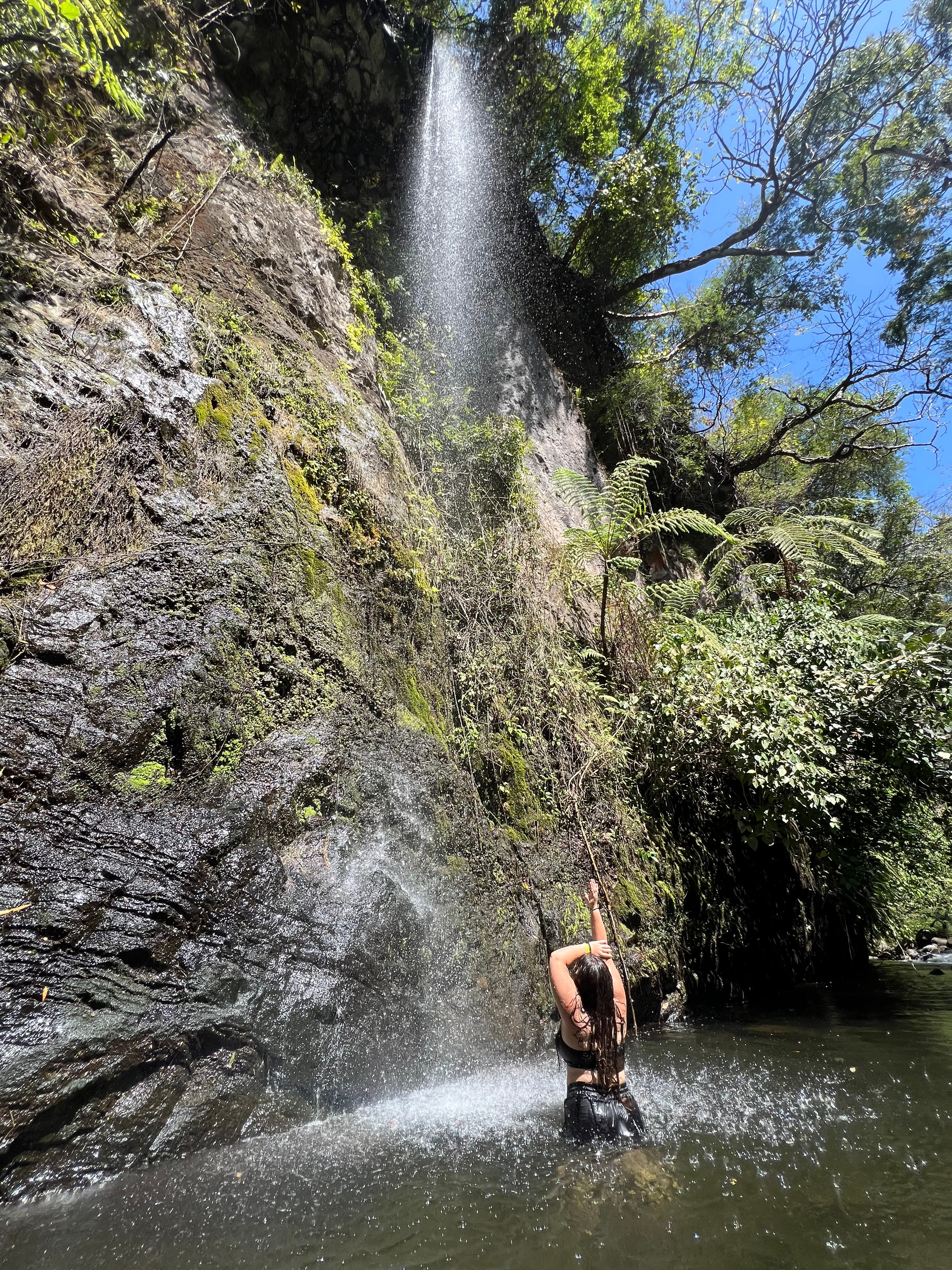 Samantha standing in a natural pool underneath a waterfall on a sunny day.