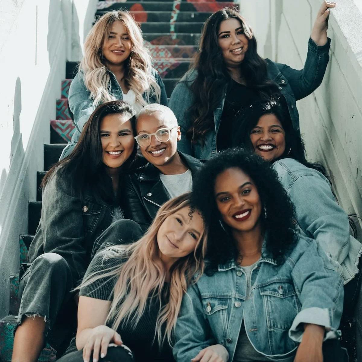 A group of seven diverse women wearing denim-focused outfits sit together in rows on an outdoor staircase, smiling at the camera and embracing each other.