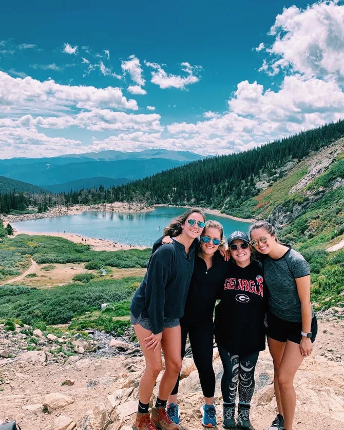 A picture of Novia with friends in hiking gear in front of a lake at the center of a mountain range with green trees and rocky terrain