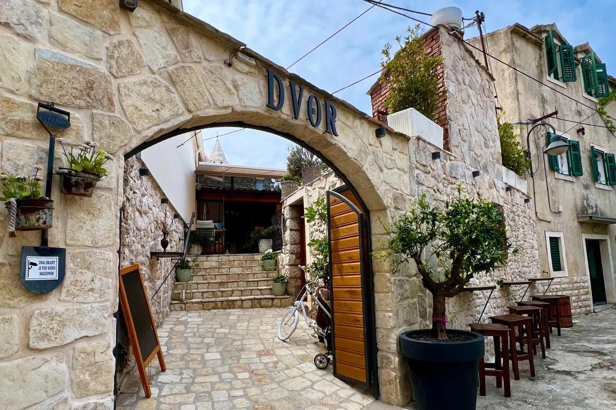 A stone archway with a metal sign saying "DVOR" alongside wooden stools, a potted tree and stone steps leading to an entrance. 