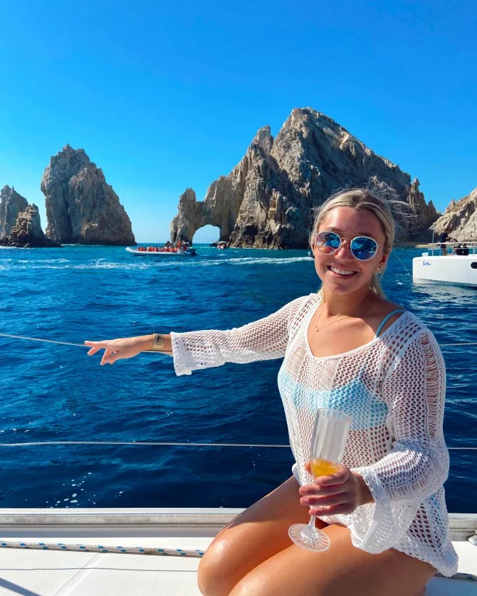 Zoe wearing a blue swimsuit and white cover-up while sitting on a boat holding a cocktail overlooking the blue water and rock formations in the distance
