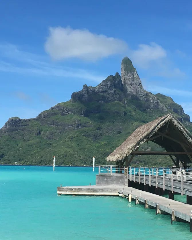 A view of Bora Bora with a wooden boardwalk, turquoise blue waters and green volcano in the background. 