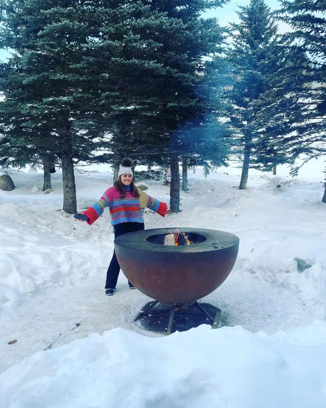 A picture of Navie in the snow standing near a round stone fire pit with pine trees in the background