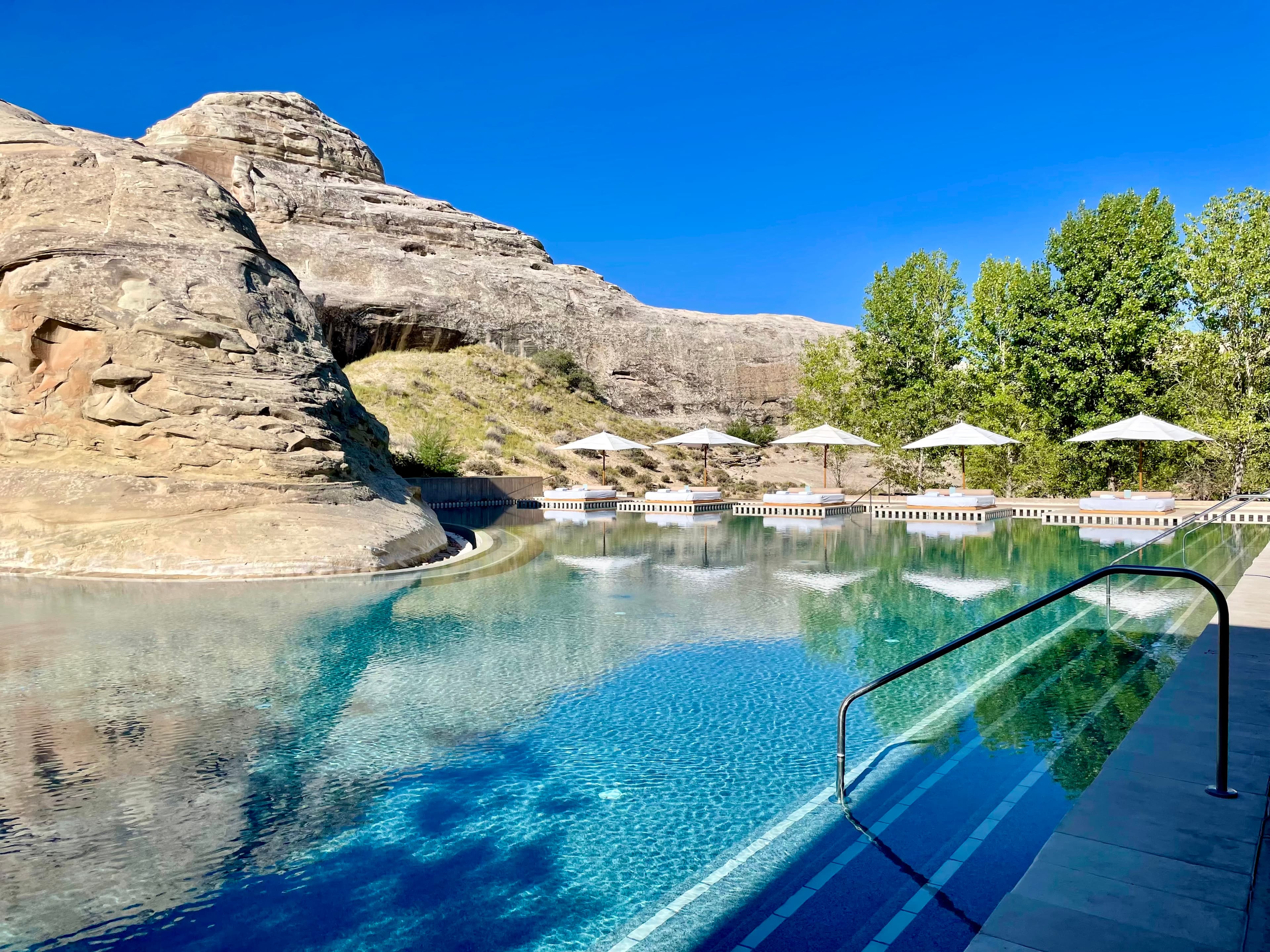 A beautiful pool with a stone wall with cabanas, trees and clear skies.
