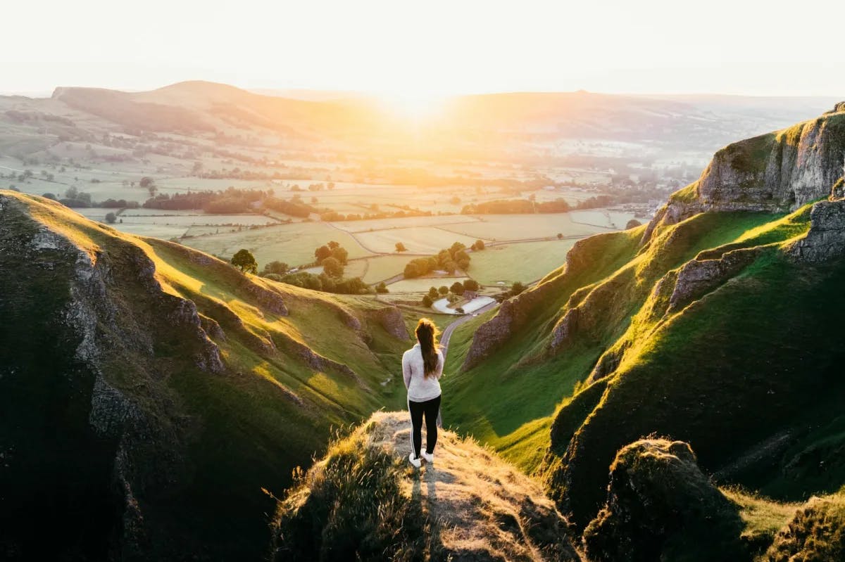 A woman stands on a hill, juxtaposed with the sun, and staring out over the green Hope Valley in the UK