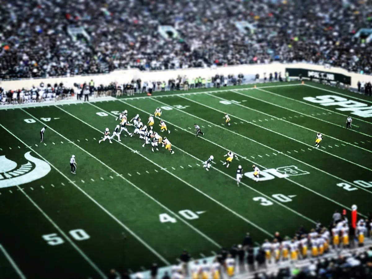 An aerial view of a large football field with two opposing teams playing against each other and crowds scattered around the stands. 