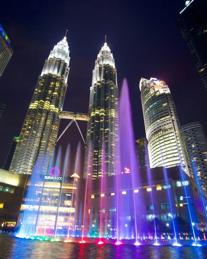 A picture of Petronas Twin Towers at night with a rainbow water fountain