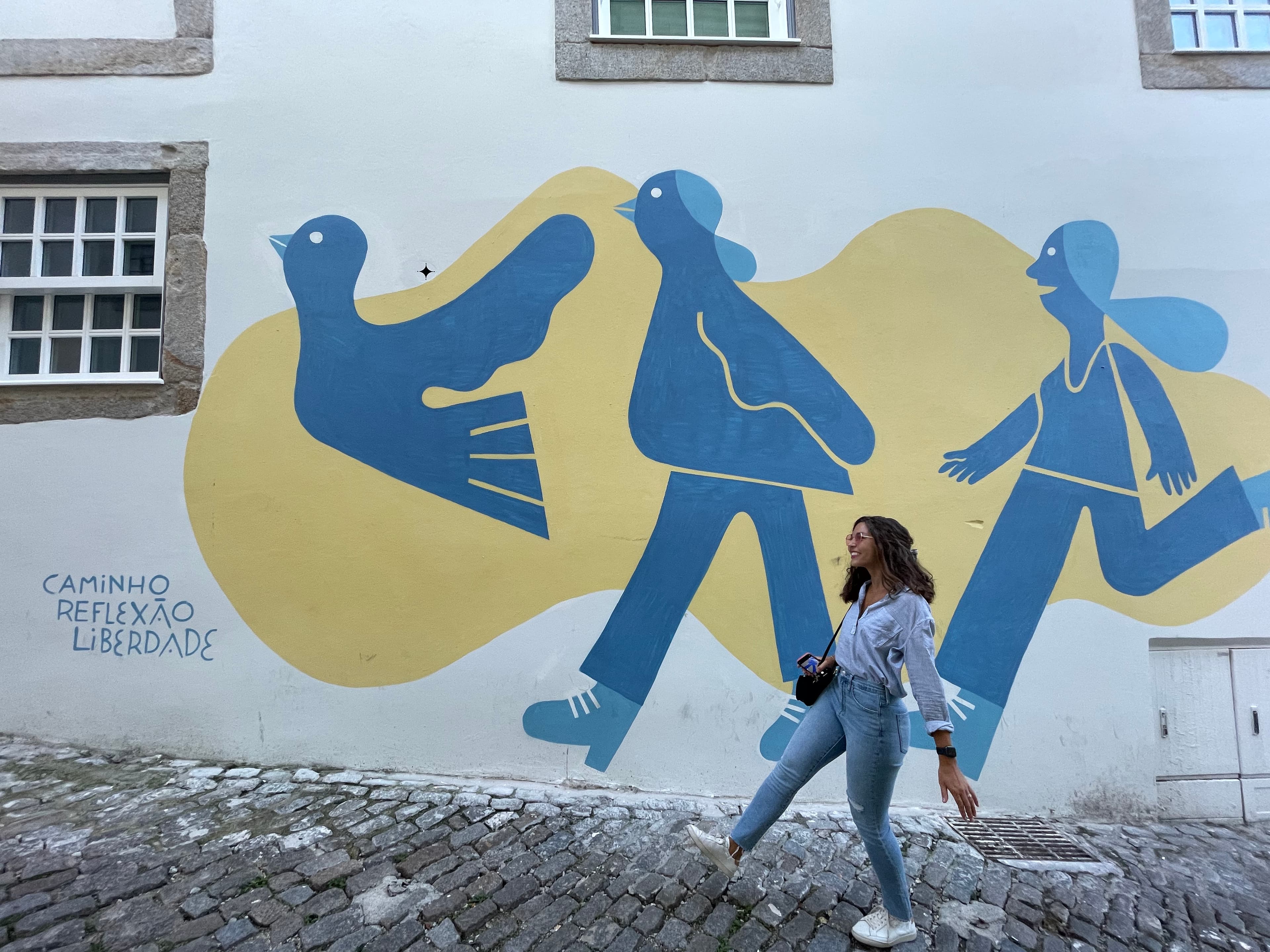 Emily posing in front of a yellow and blue mural against a white building and brick road.
