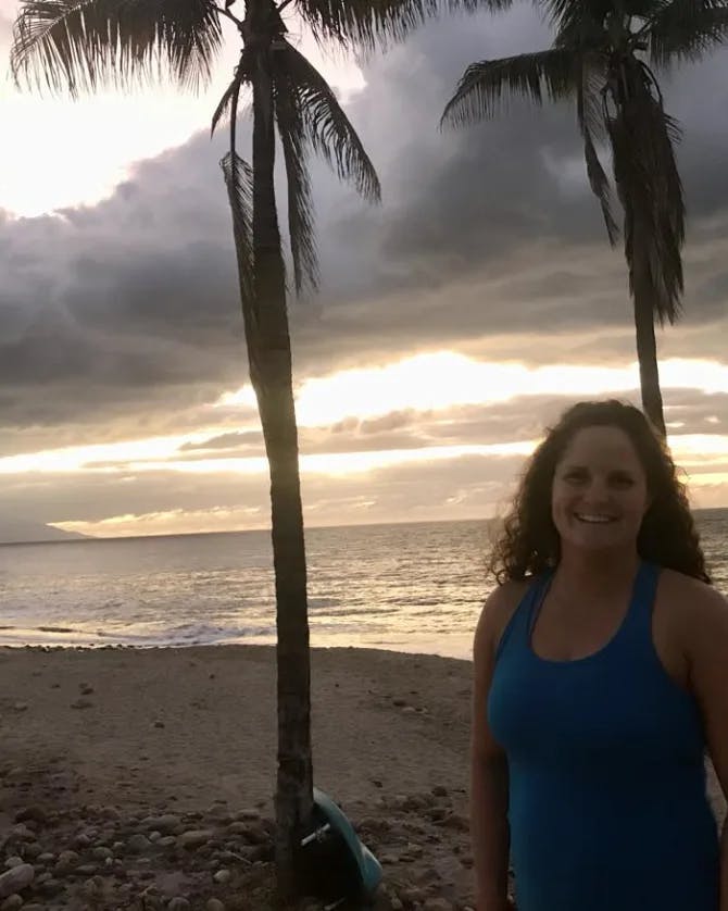 Katie wearing a blue tank top on the beach at sunset with two palm trees in the background