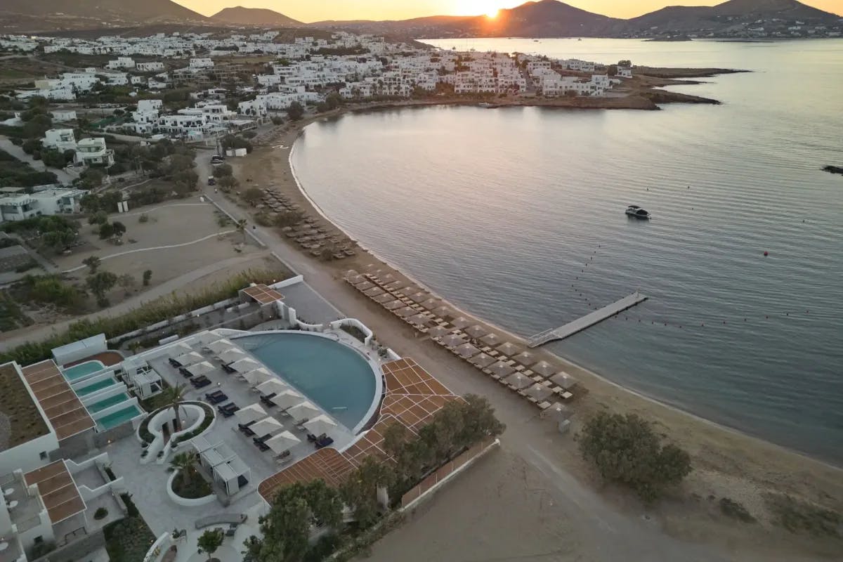 Aerial view: the sun sets over distant mountains, with loungers lining the shoreline in front of Cosme, a Luxury Collection Resort, Paros